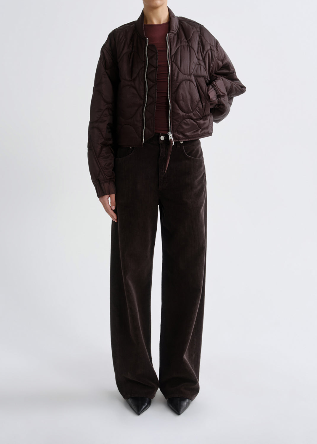 A model wearing a dark brown quilted bomber jacket, matching wide-leg corduroy trousers, a burgundy top, and black pointed shoes, photographed against a grey background.