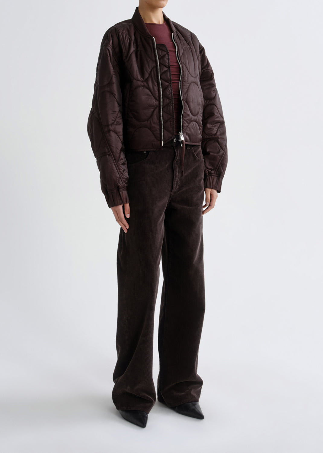 A model wearing a dark brown quilted bomber jacket, matching dark brown wide-leg corduroy trousers, and a burgundy top, standing against a grey background.