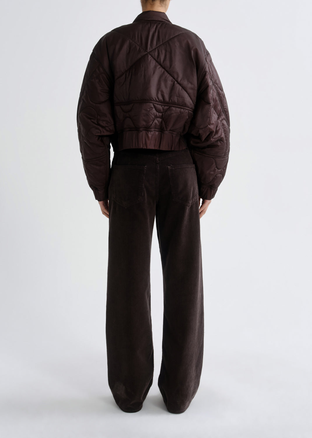 A model shown from the back wearing a dark brown quilted bomber jacket and matching dark brown wide-leg trousers against a grey background.