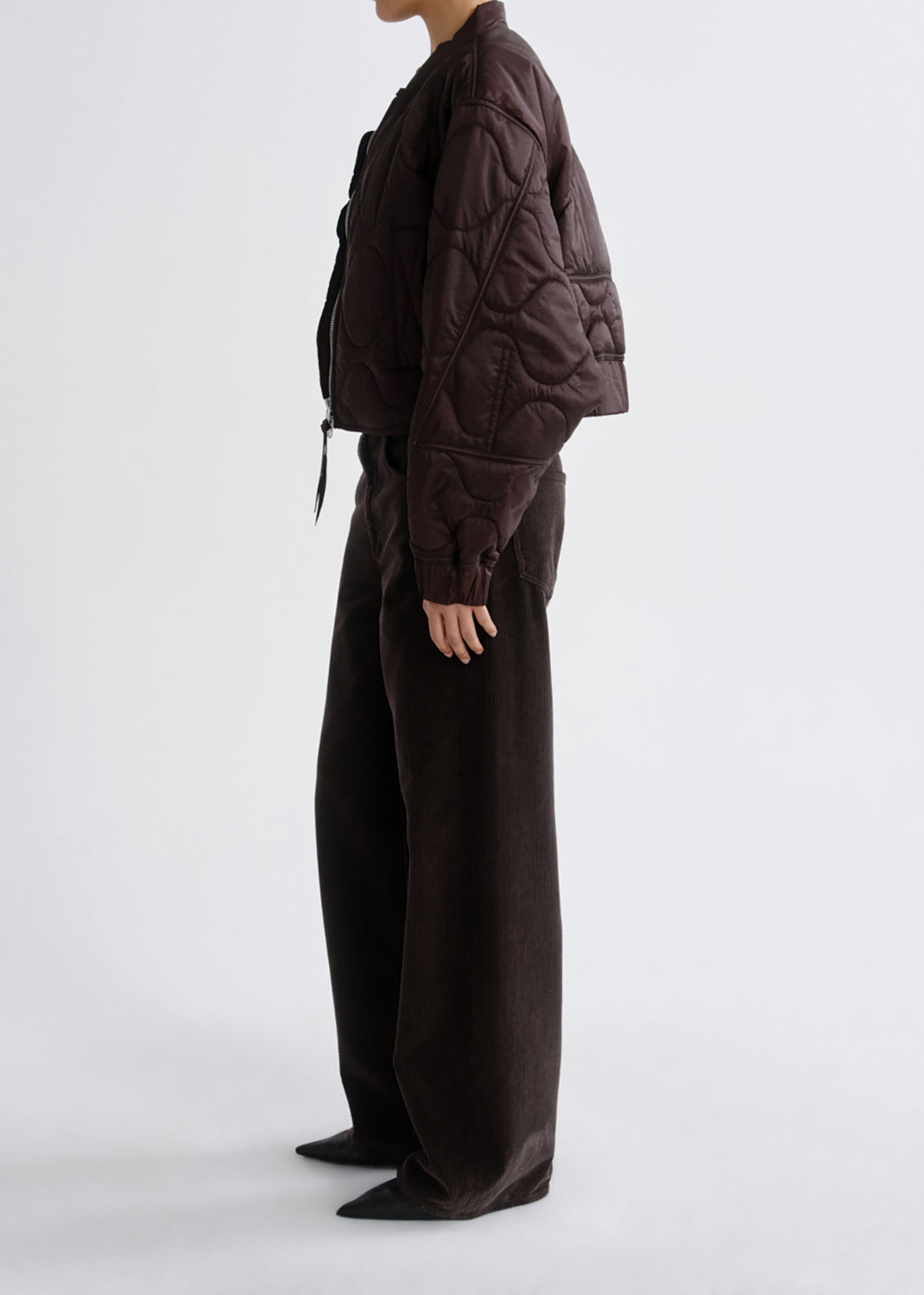 A model shown from the side wearing a dark brown quilted cropped jacket and matching wide-leg trousers against a grey background.