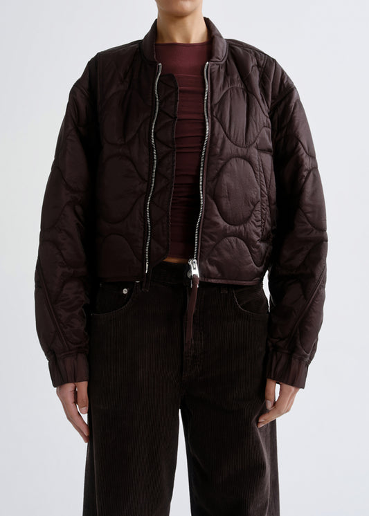 A model wearing a dark brown quilted bomber jacket over a dark burgundy top and dark brown corduroy pants, shown from the neck down against a grey background.