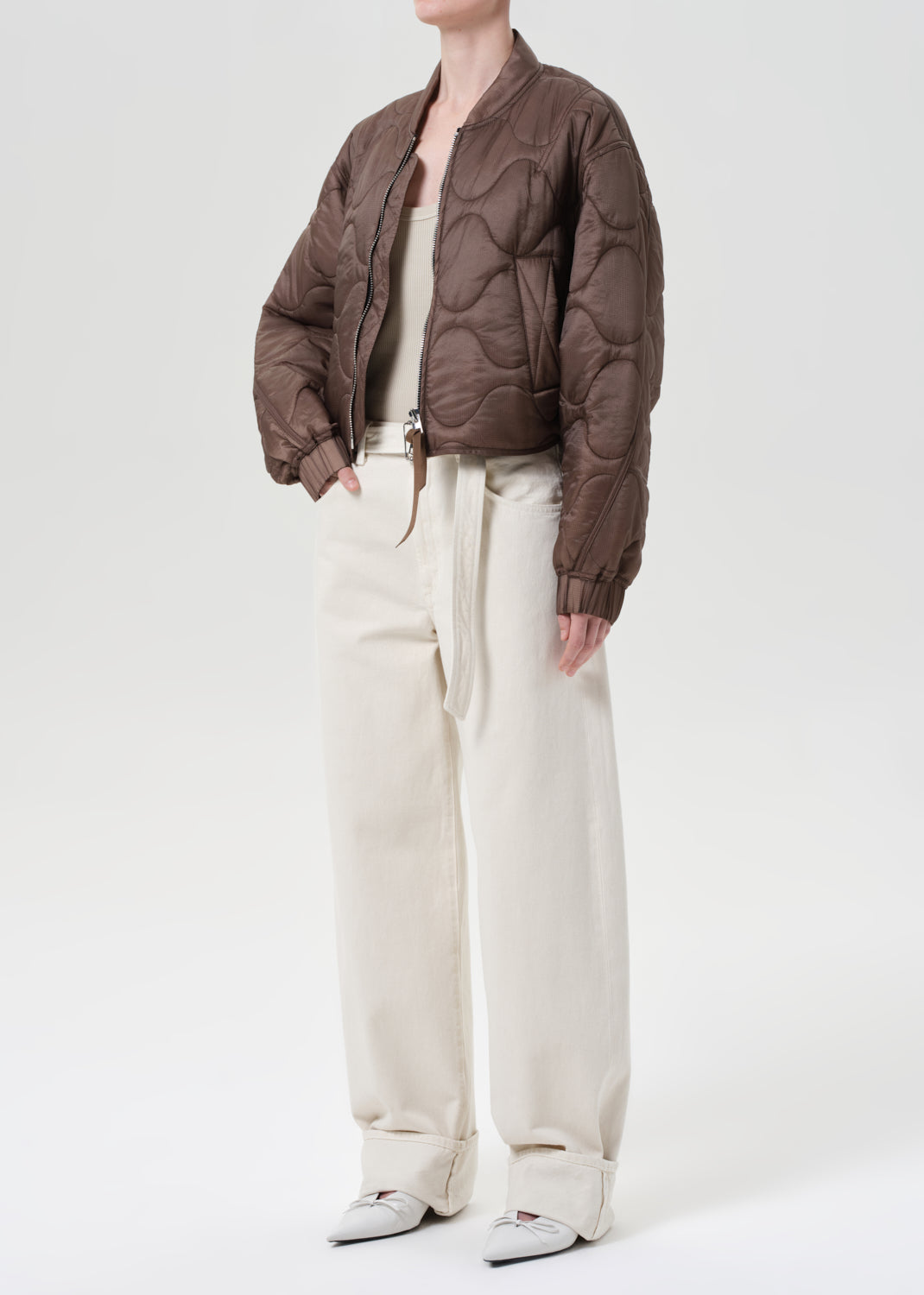Person wearing a brown quilted jacket and white pants on a white background