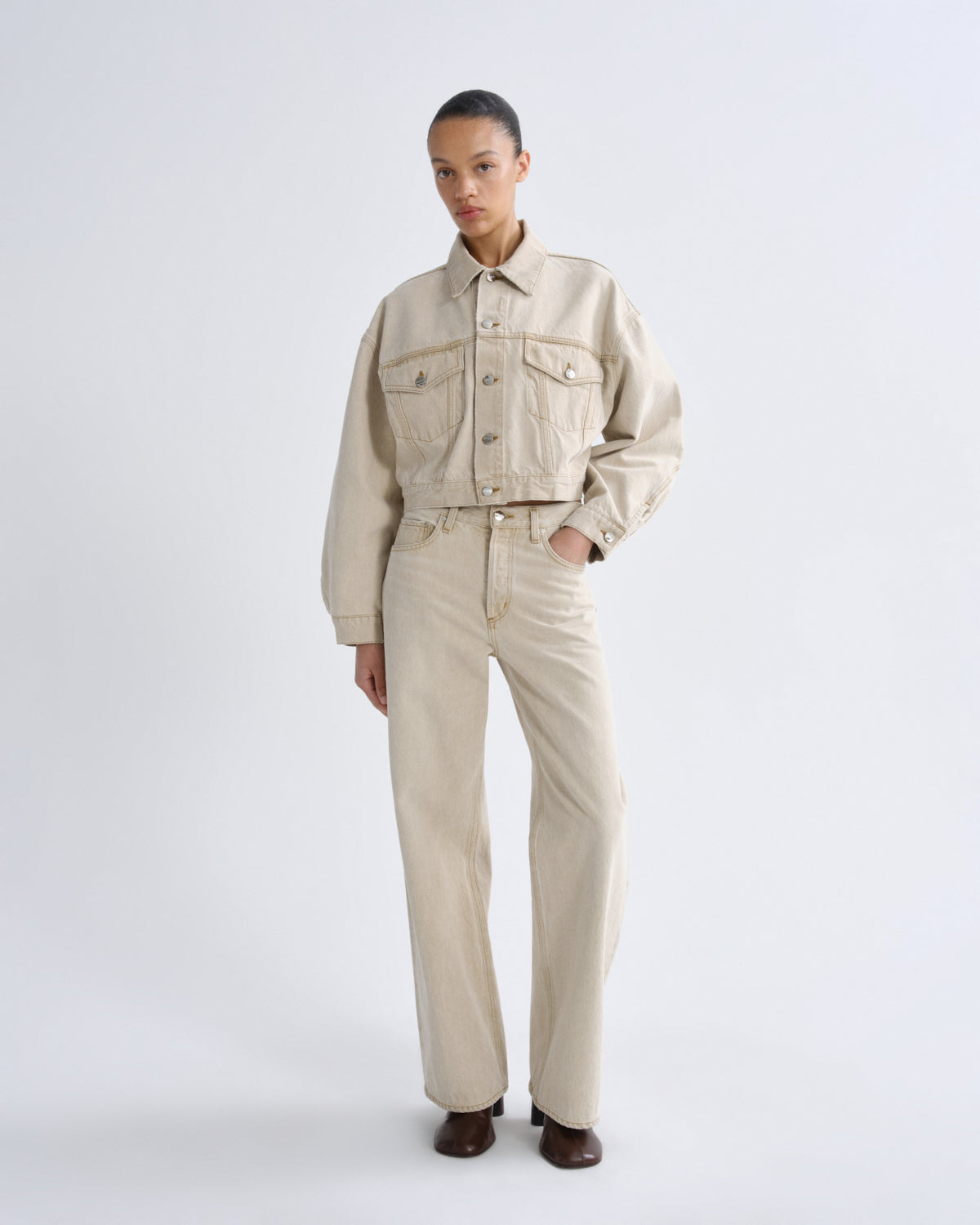 A model stands against a light gray studio background wearing a light beige cropped denim jacket with front flap pockets and matching straight-leg denim pants, paired with dark brown shoes.