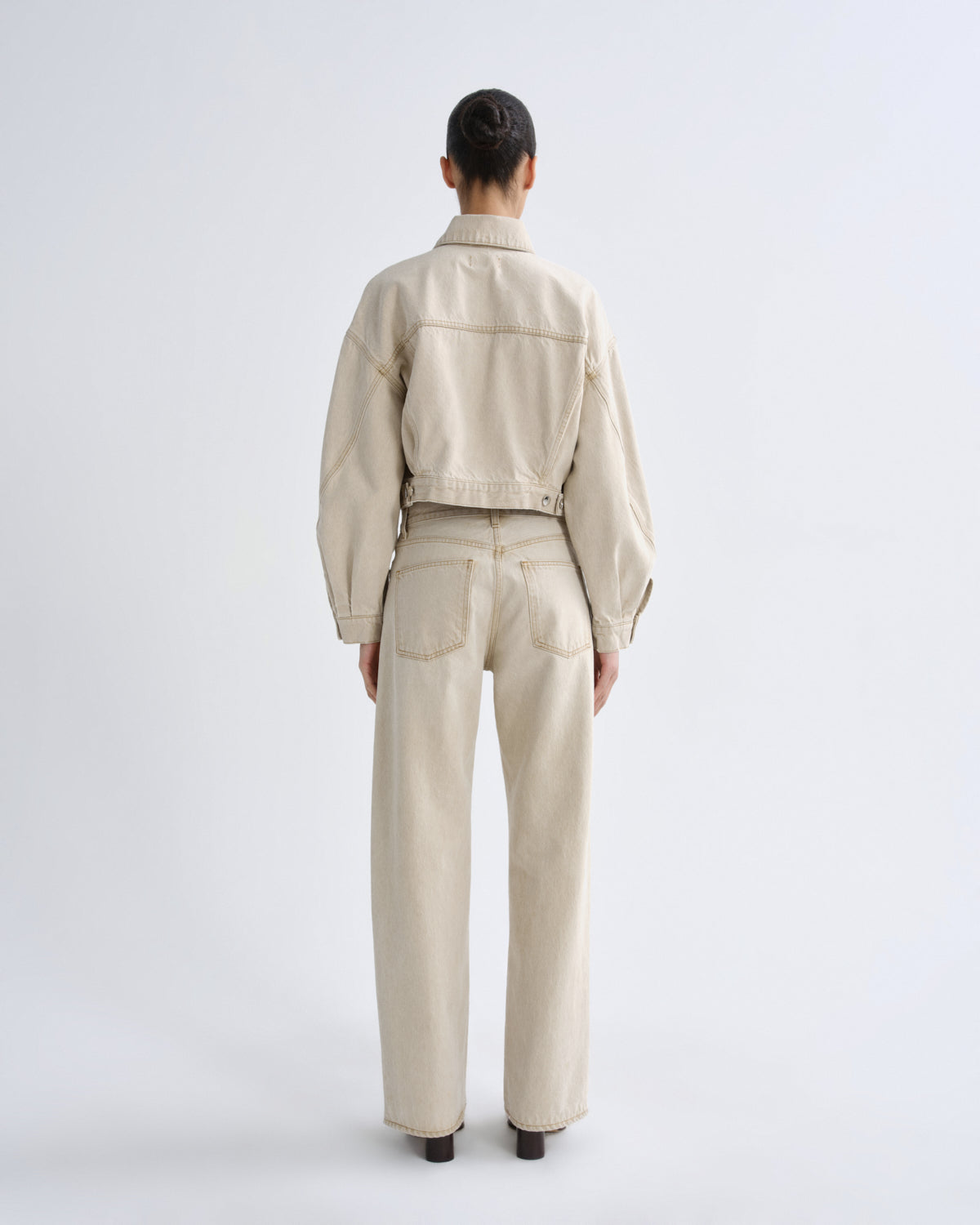 A model viewed from the back wearing a light beige cropped jacket and matching wide-leg trousers against a plain light gray studio background.