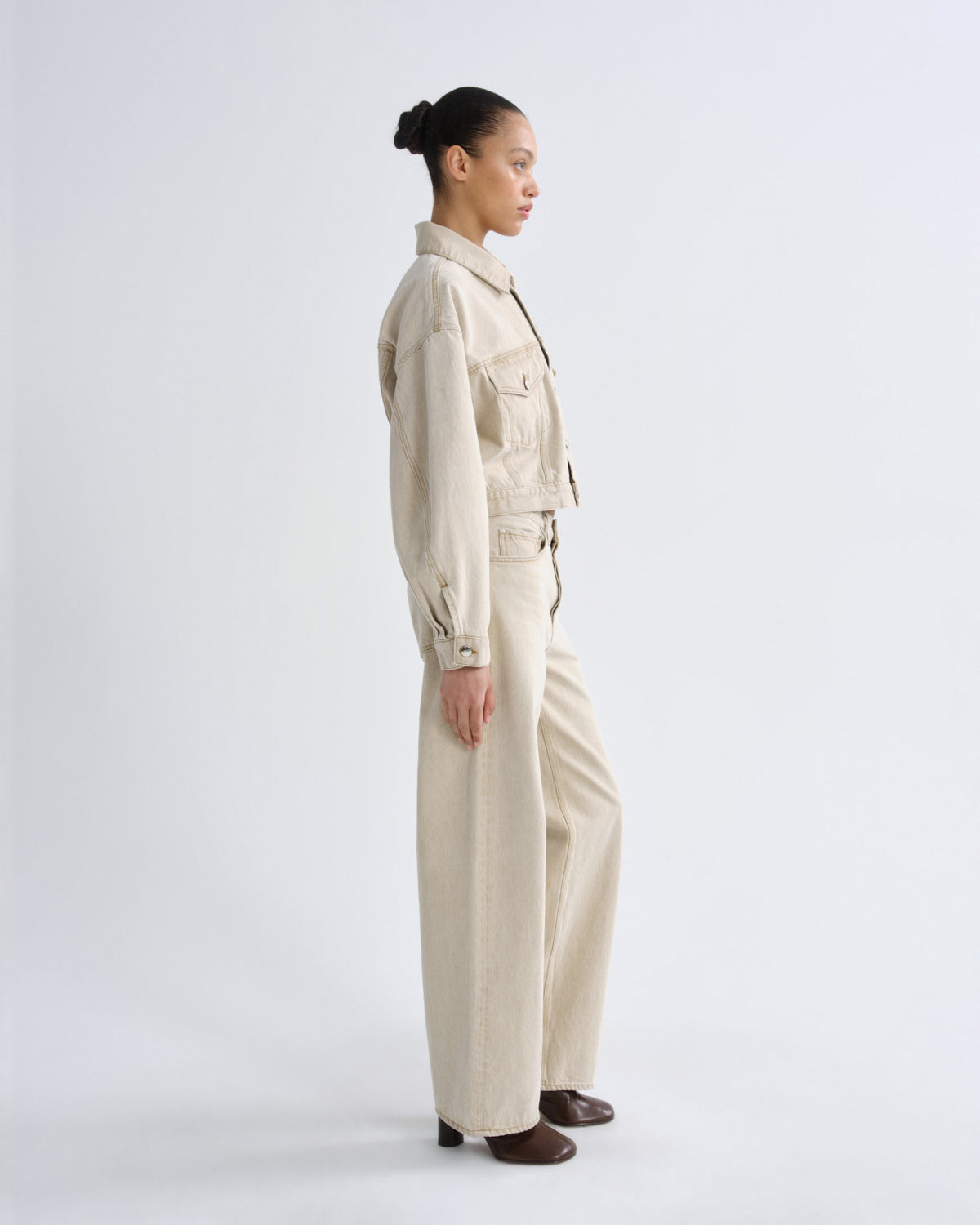 A model in profile wearing a light beige utility jacket and matching wide-leg trousers, styled with dark brown shoes against a plain light gray studio background.