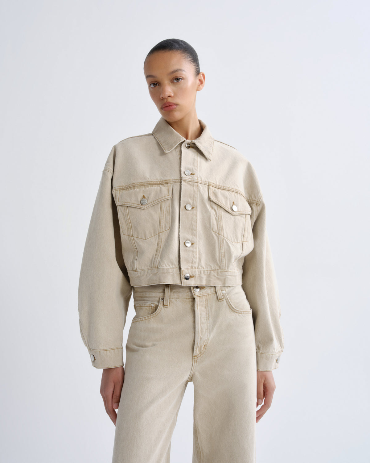 A model wearing a light beige cropped denim jacket with front flap pockets and matching beige wide-leg jeans, photographed against a clean light gray studio background.