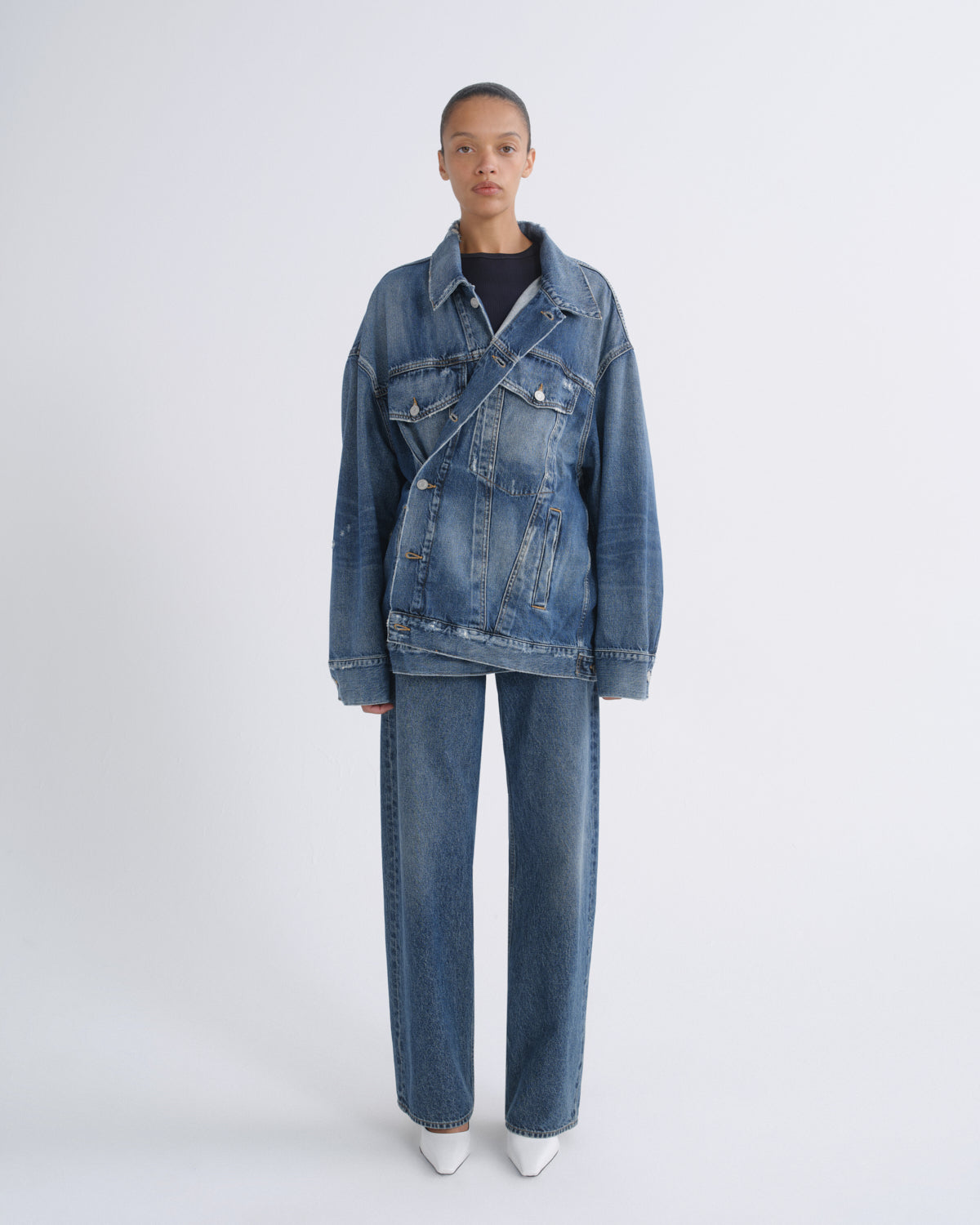 A model stands against a light gray studio background wearing a blue denim jacket and matching wide-leg jeans over a black top and white pointed shoes.