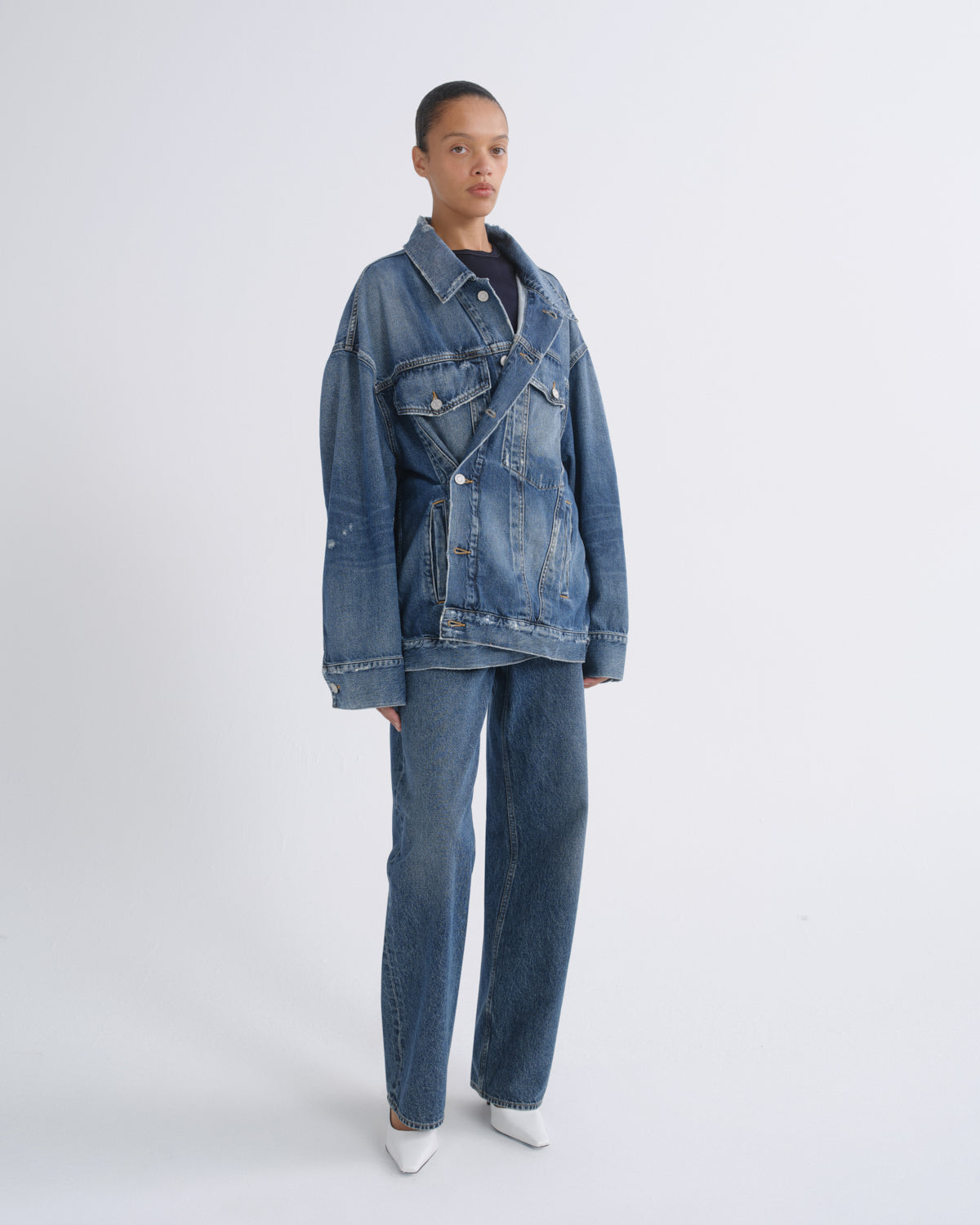 A model stands against a light gray studio background wearing a blue denim jacket and matching wide-leg jeans over a dark top, with white pointed shoes.