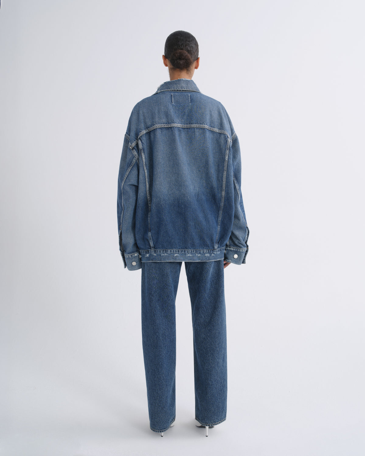 A model viewed from the back wearing an oversized blue denim jacket and matching wide-leg denim jeans against a plain light gray studio background.