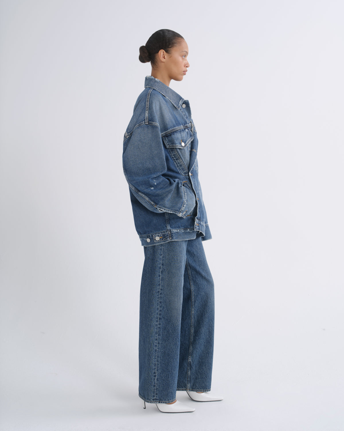 A model shown in profile wearing a blue denim jacket and matching wide-leg jeans, standing against a plain light gray studio background.