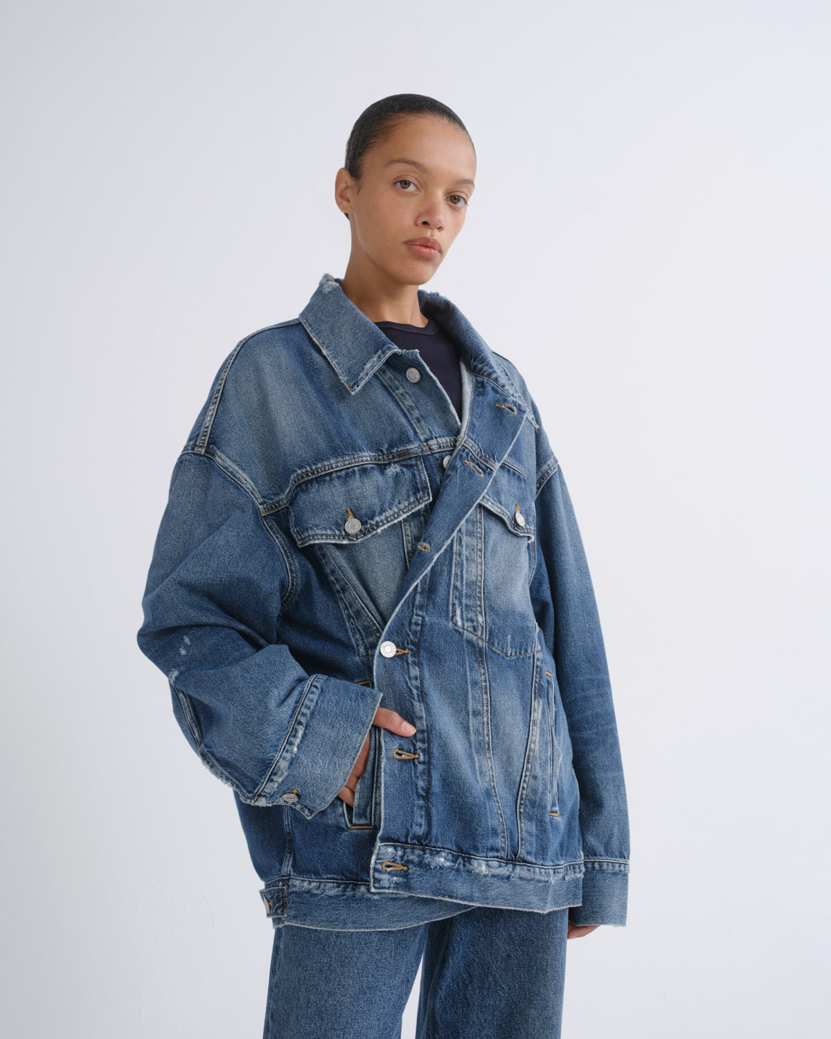 A model wearing an oversized blue denim jacket with a buttoned wrap-style front, worn over a dark top and paired with matching blue jeans, photographed against a light gray studio background.