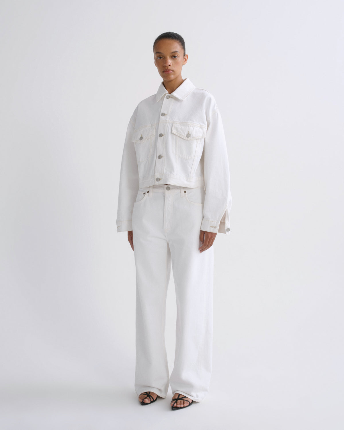 A model stands against a plain light gray studio background wearing a matching white denim outfit consisting of a cropped button-front jacket and wide-leg pants, paired with black sandals.
