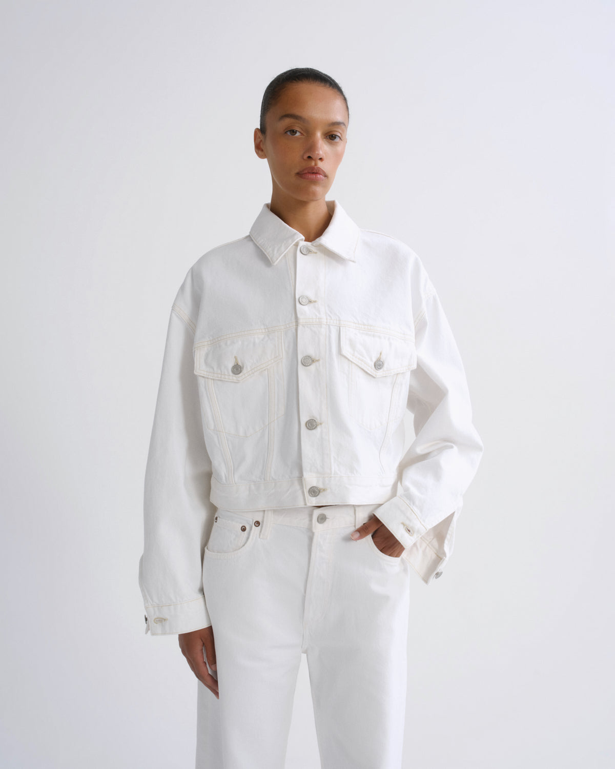 A model wearing a white cropped denim jacket with silver buttons and white jeans, standing against a plain light gray studio background.