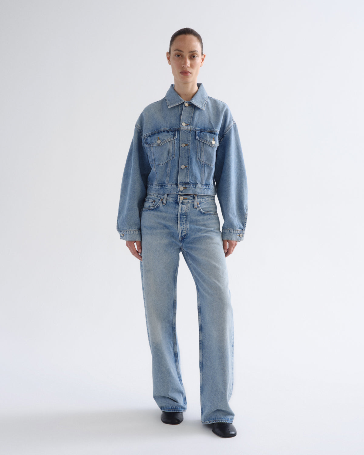 A model stands against a light gray studio background wearing a light blue denim jacket and matching wide-leg denim jeans.