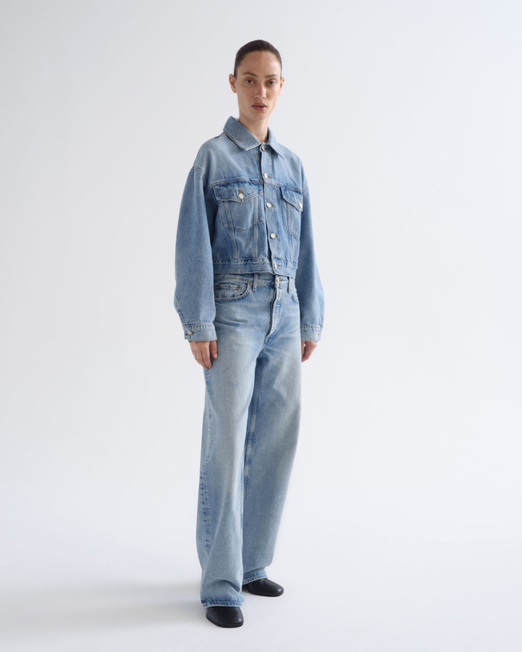 A model stands against a plain light gray studio background wearing a light-wash blue denim jacket and matching wide-leg denim jeans.
