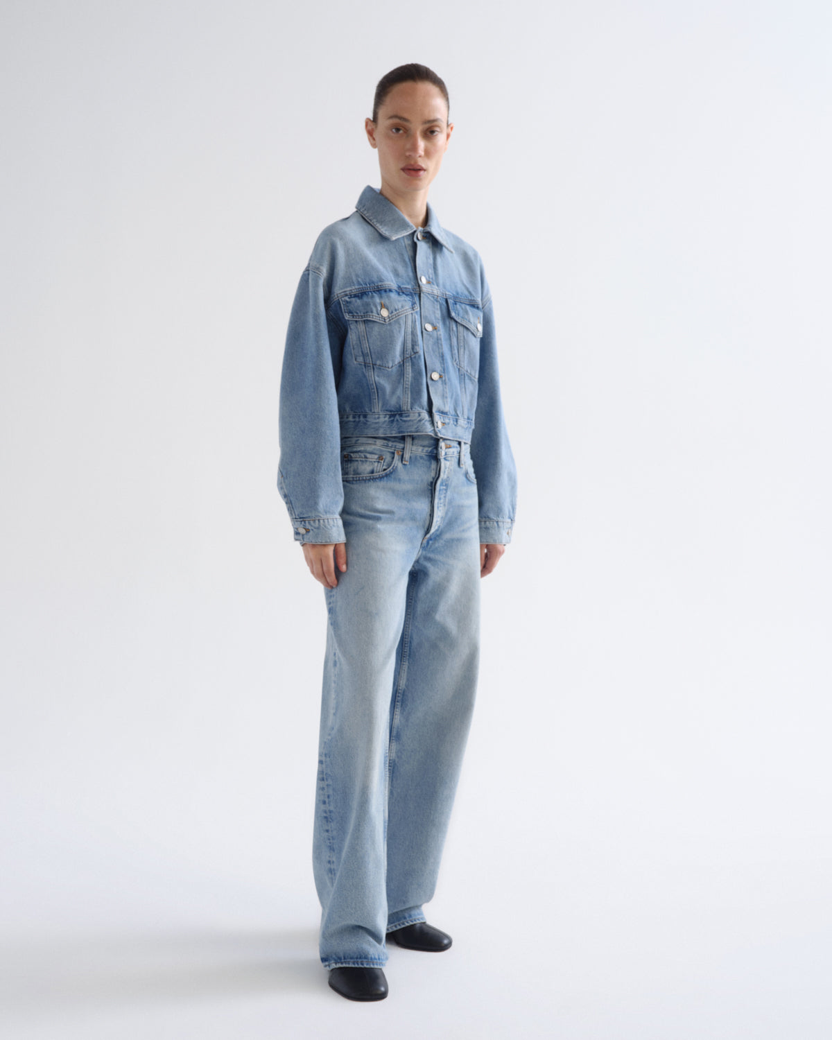 A model stands against a plain light gray studio background wearing a light-wash blue denim jacket and matching wide-leg denim jeans.