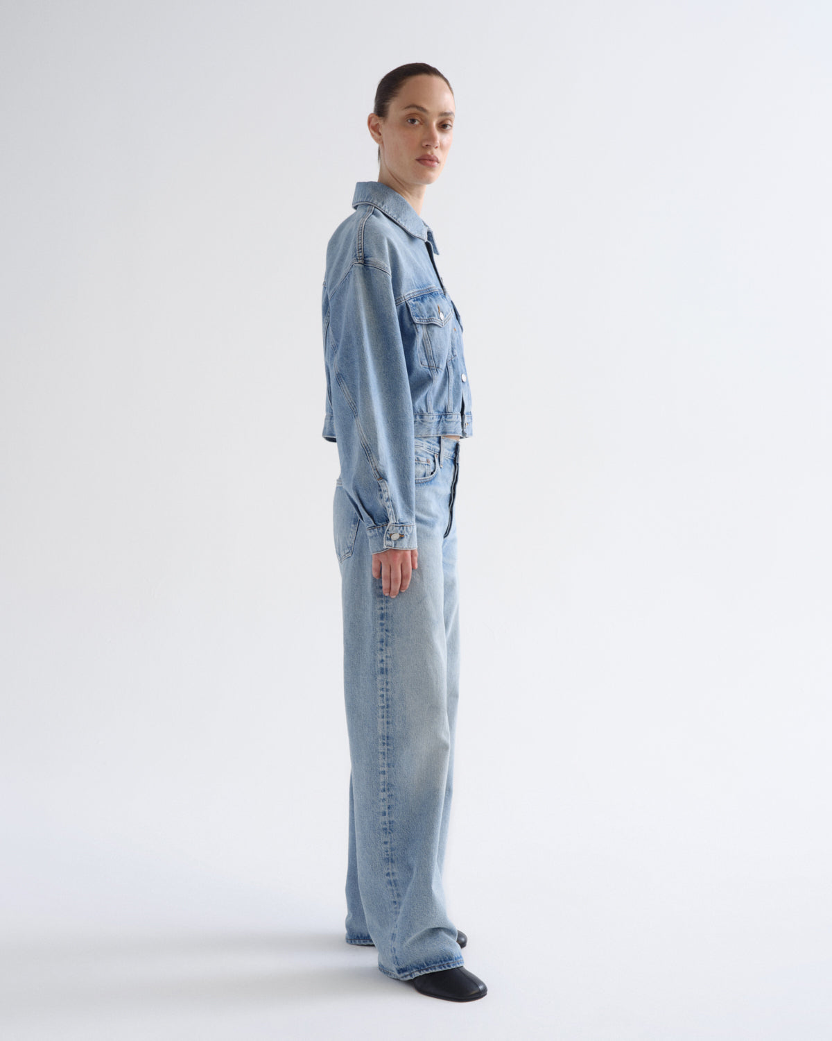 A model stands in profile wearing a light blue denim jumpsuit with a button-up shirt collar and chest pockets, paired with black slip-on shoes, against a plain white studio background.