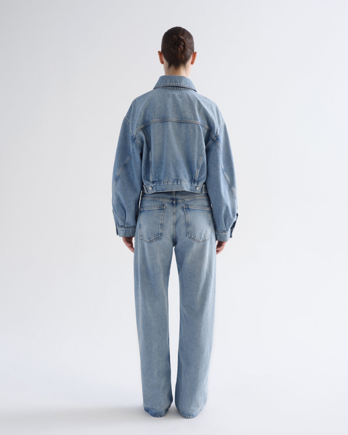A model viewed from the back wearing a light-wash blue denim jacket and matching wide-leg denim jeans against a plain light gray studio background.