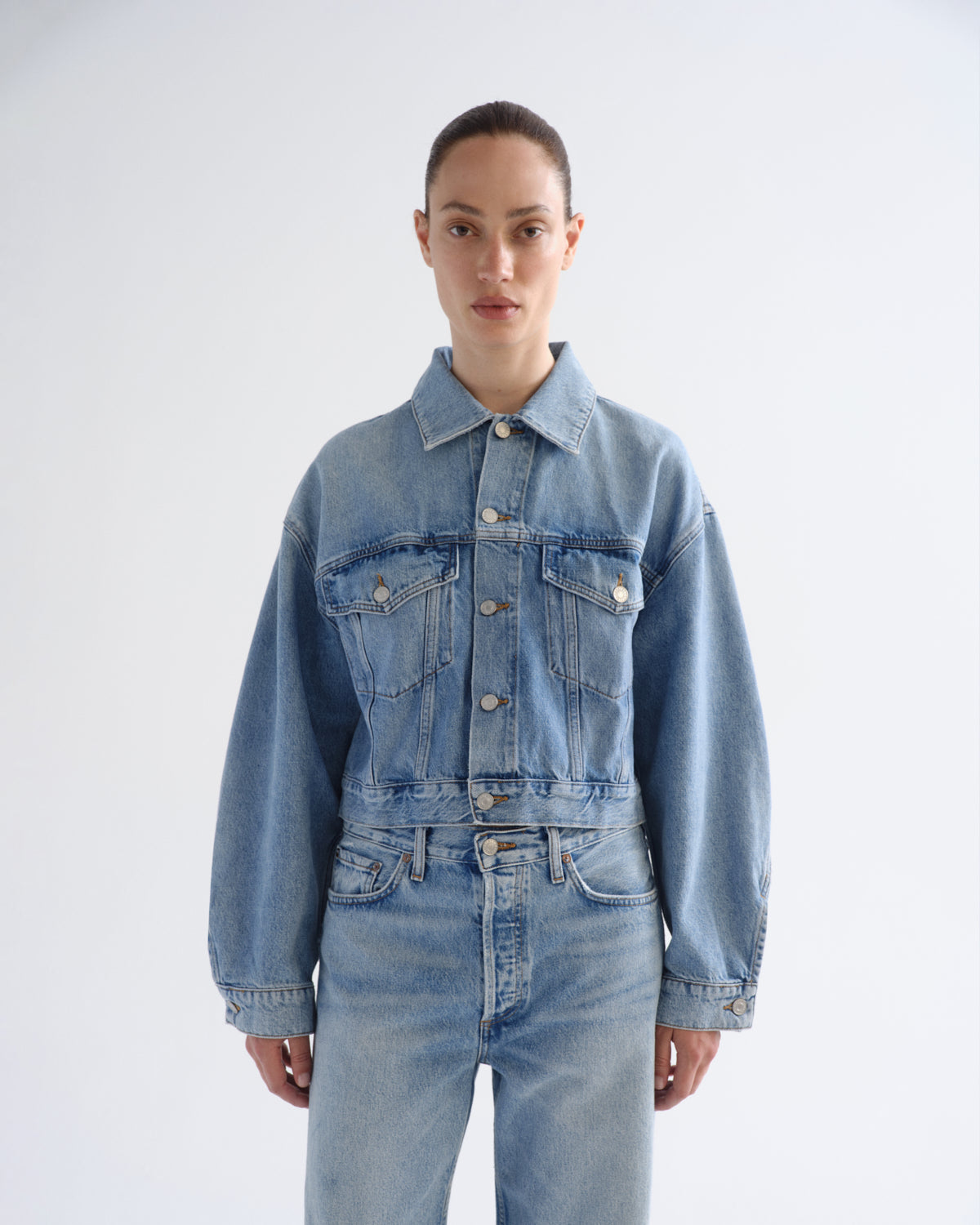 A model wearing a light blue cropped denim jacket with matching light blue jeans, standing against a plain light gray studio background.