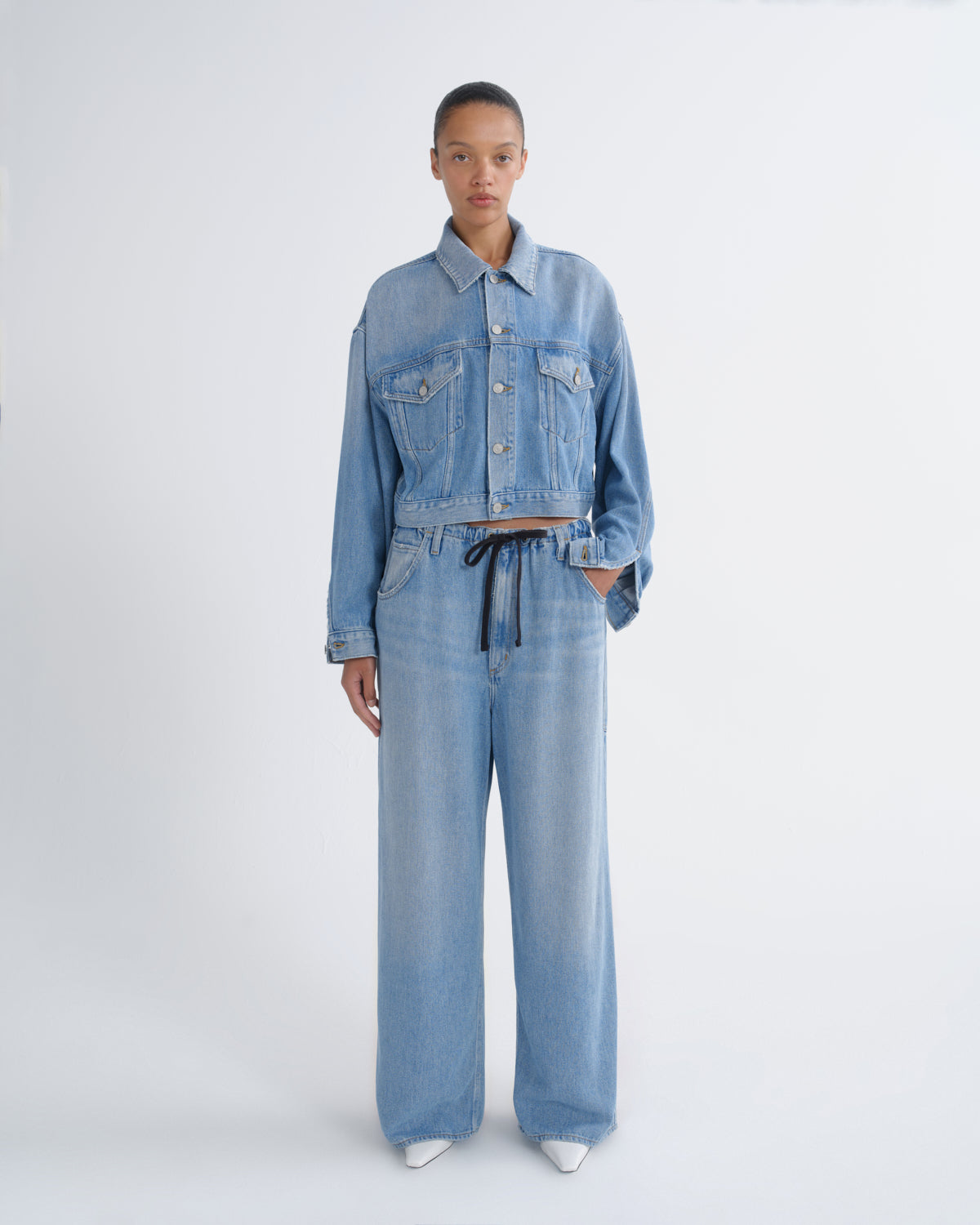 A model stands against a light gray studio background wearing a light blue denim set consisting of a cropped button-front jacket with long sleeves and matching wide-leg pants with a dark drawstring waist.