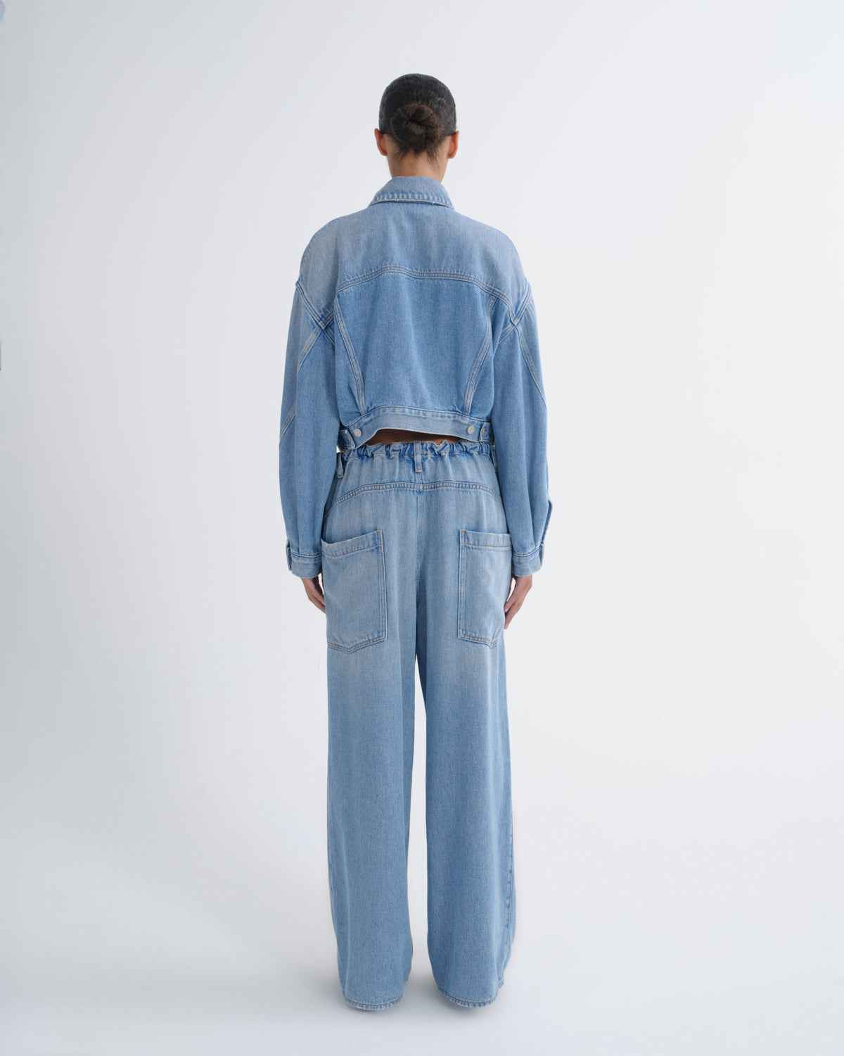 A model viewed from the back wearing a light blue denim jumpsuit with wide-leg pants, a cropped long-sleeve top, and a matching belt.