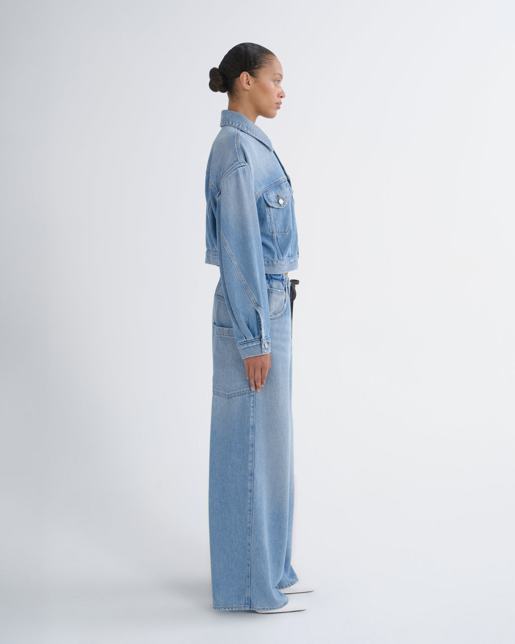 A model in side profile wearing a light blue denim set consisting of a cropped button-up jacket and matching wide-leg trousers, photographed against a seamless light gray studio background.
