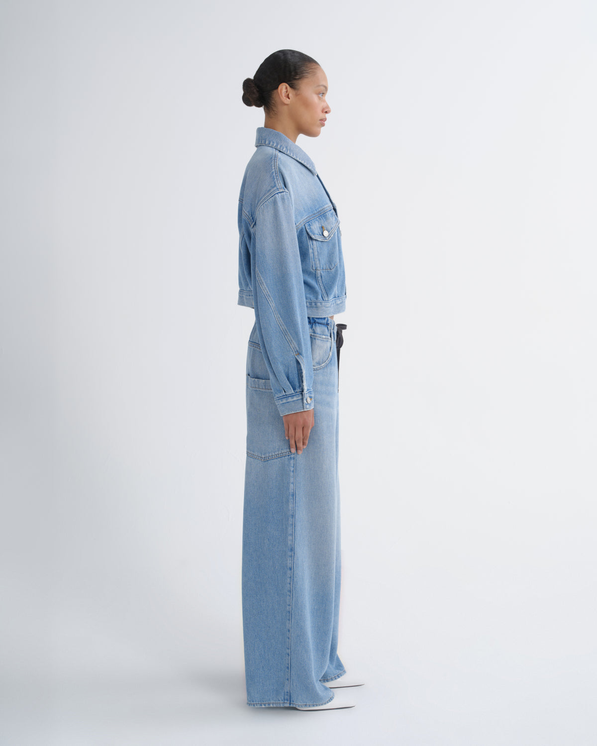 A model in side profile wearing a light blue denim set consisting of a cropped button-up jacket and matching wide-leg trousers, photographed against a seamless light gray studio background.