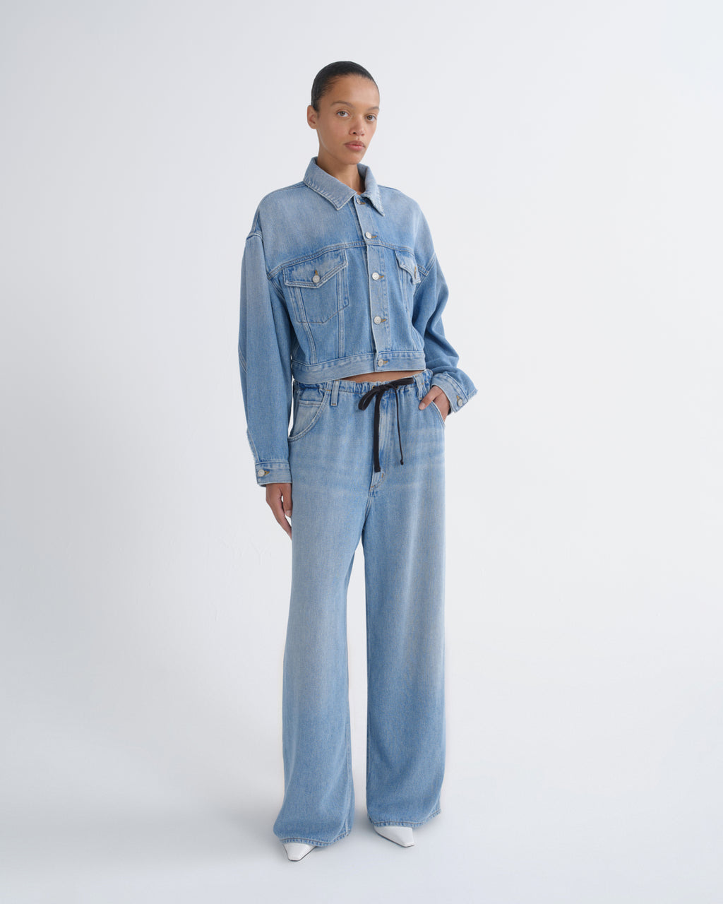 A model stands against a light gray studio background wearing a light blue denim set consisting of a cropped button-front jacket and matching wide-leg pants with a dark drawstring.