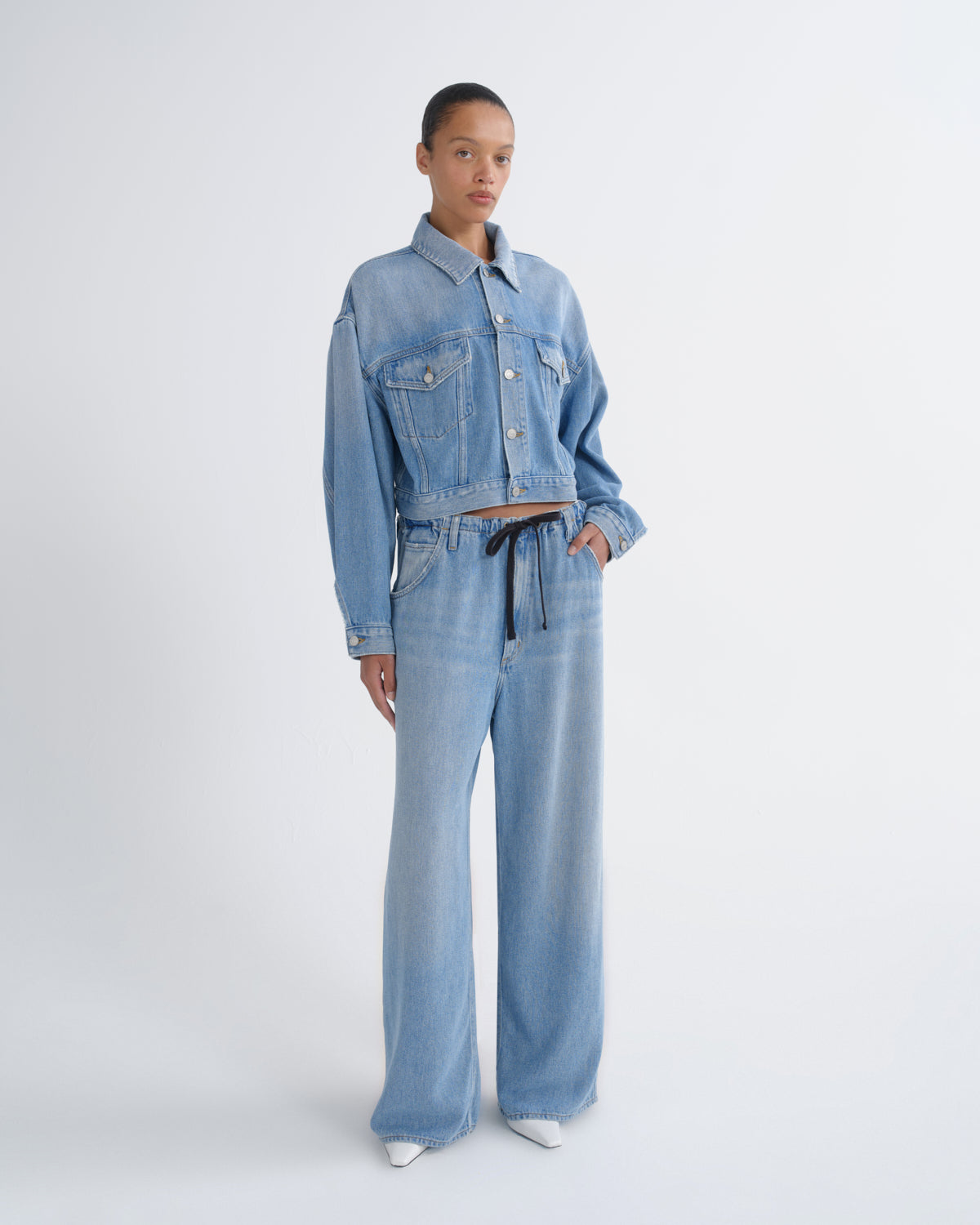 A model stands against a light gray studio background wearing a light blue denim set consisting of a cropped button-front jacket and matching wide-leg pants with a dark drawstring.