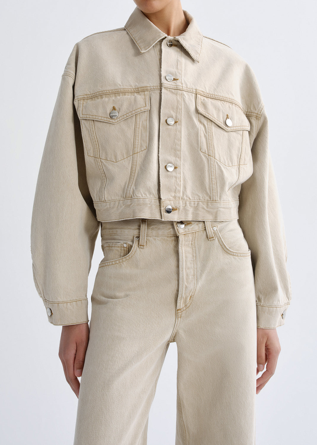 Person wearing a beige denim jacket and jeans on a grey background