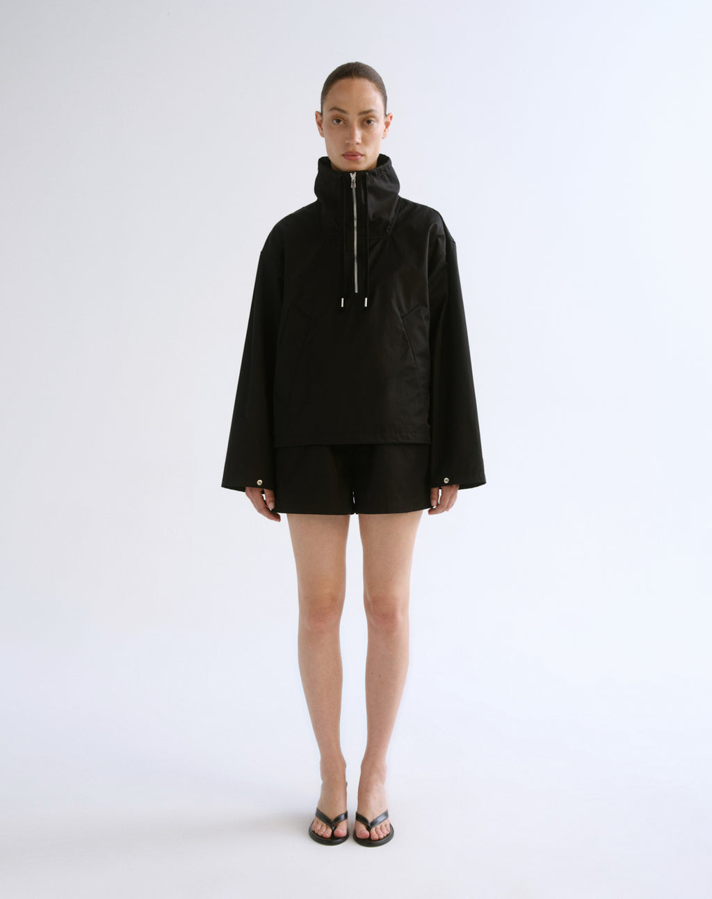 A model stands against a plain light gray studio background wearing a black hooded zip-front jacket with long sleeves, black shorts, and black sandals.