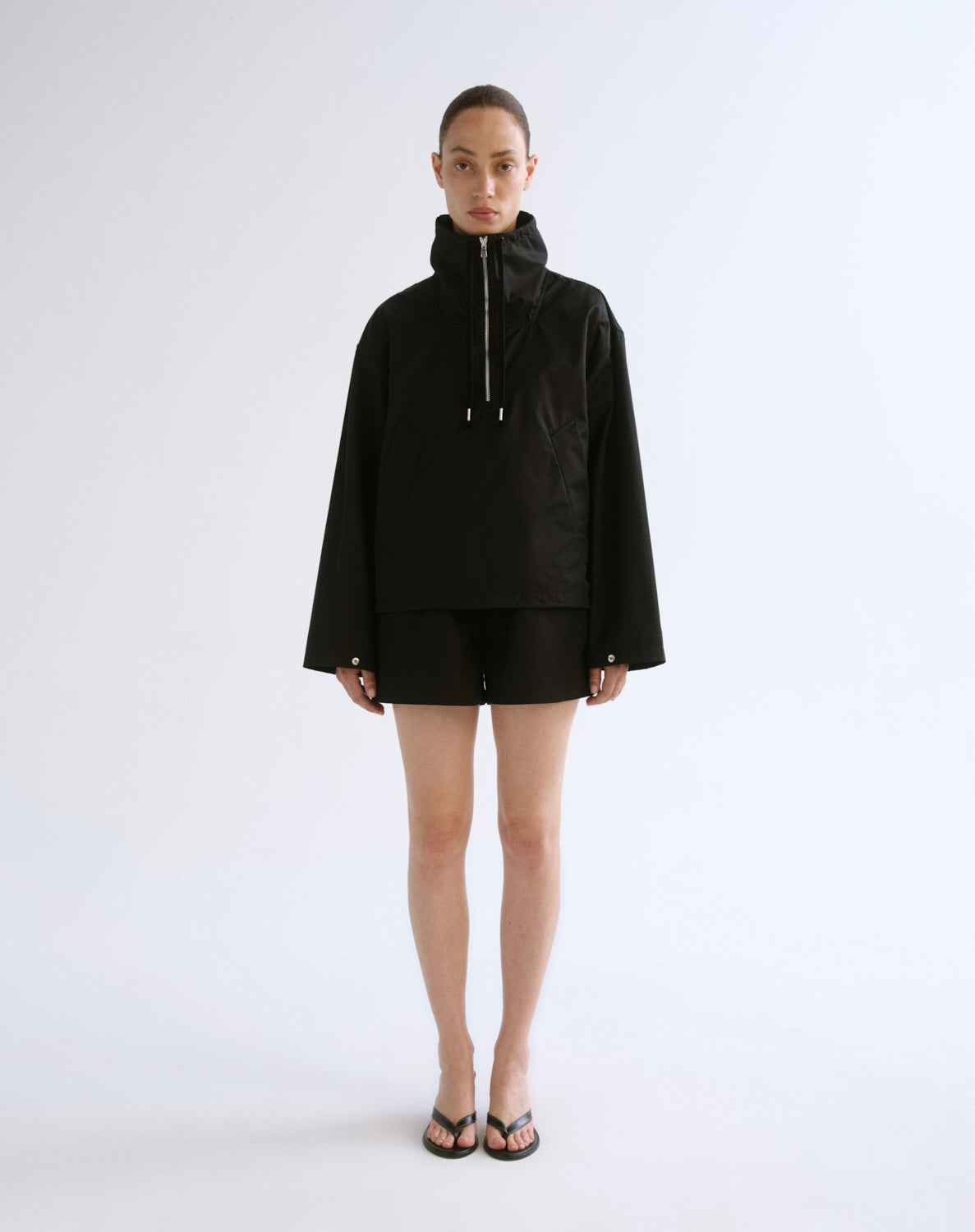 A model stands against a plain light gray studio background wearing a black hooded zip-front jacket with long sleeves, black shorts, and black sandals.
