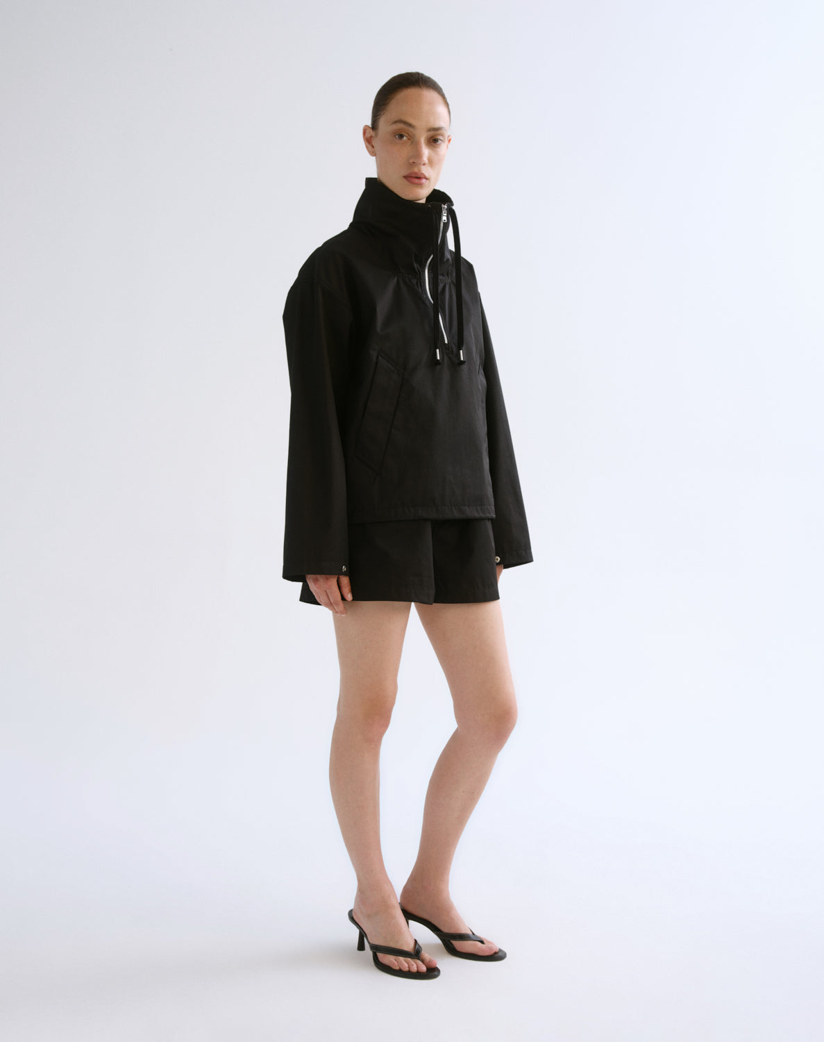 A model stands against a light gray studio background wearing a black hooded jacket with a high collar, black shorts, and black heeled sandals.