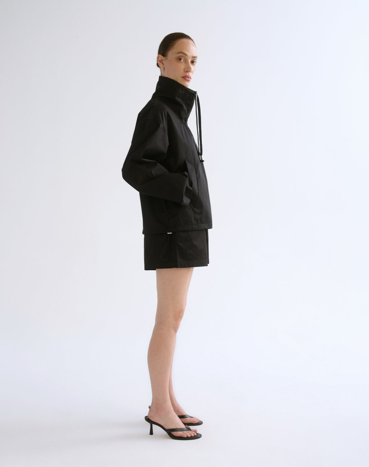 A model stands in profile wearing a black high-neck jacket with a drawstring, a matching black mini skirt, and black heeled sandals against a plain light gray studio background.