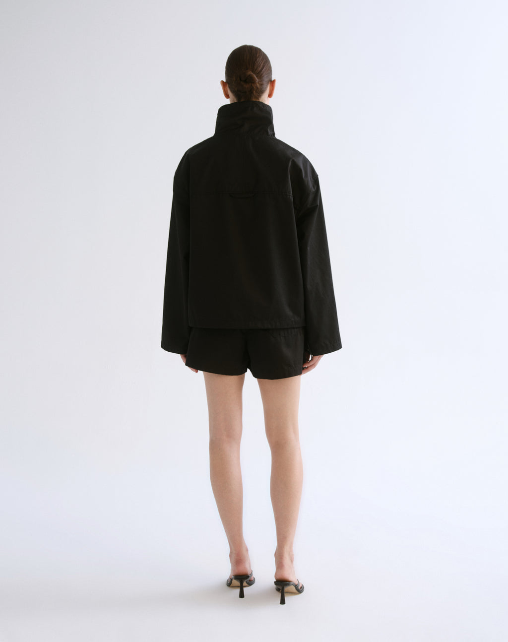 A model viewed from the back wearing a black high-neck jacket and matching black shorts, paired with black pointed-toe heeled sandals, against a plain light gray studio background.