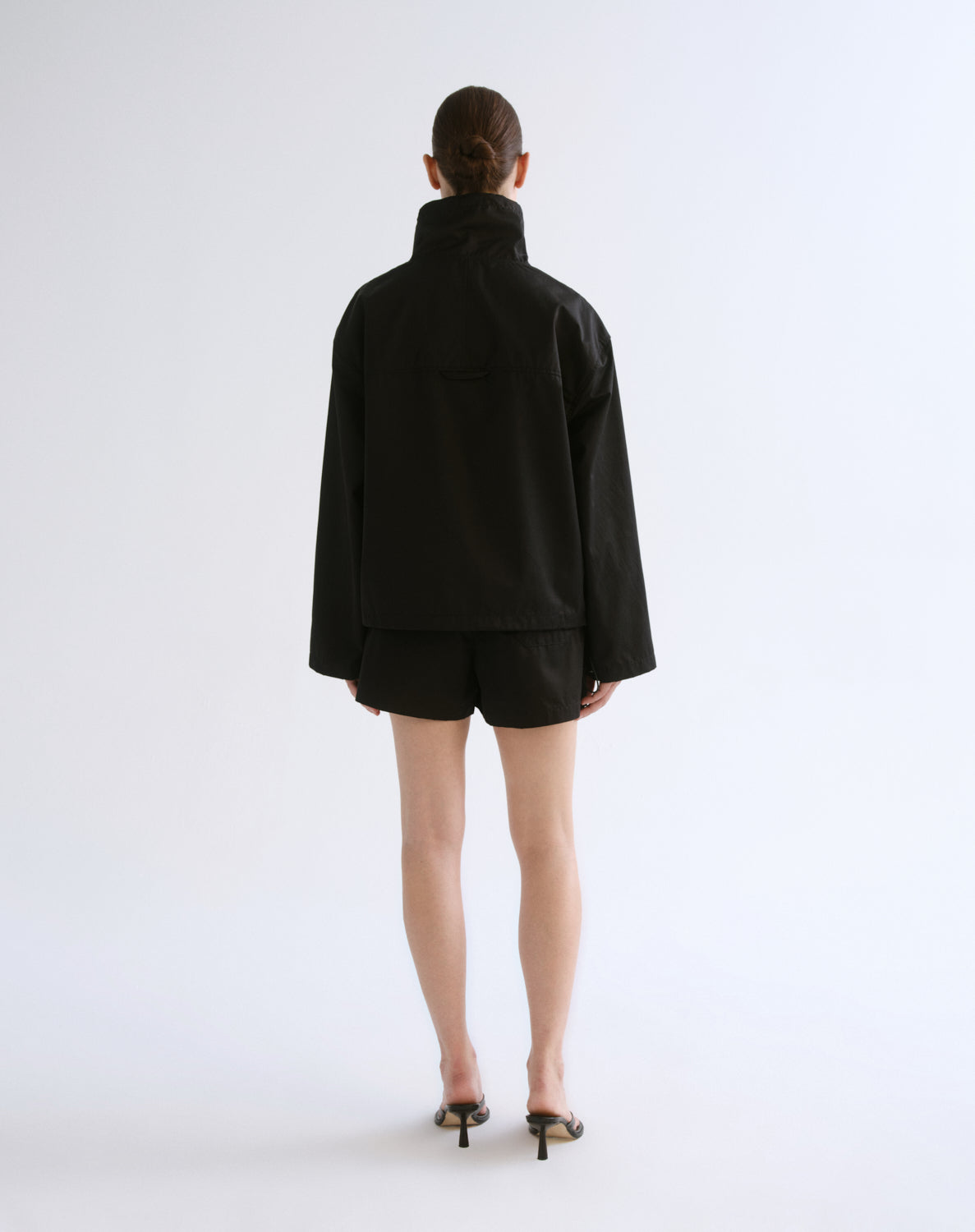 A model viewed from the back wearing a black high-neck jacket and matching black shorts, paired with black pointed-toe heeled sandals, against a plain light gray studio background.