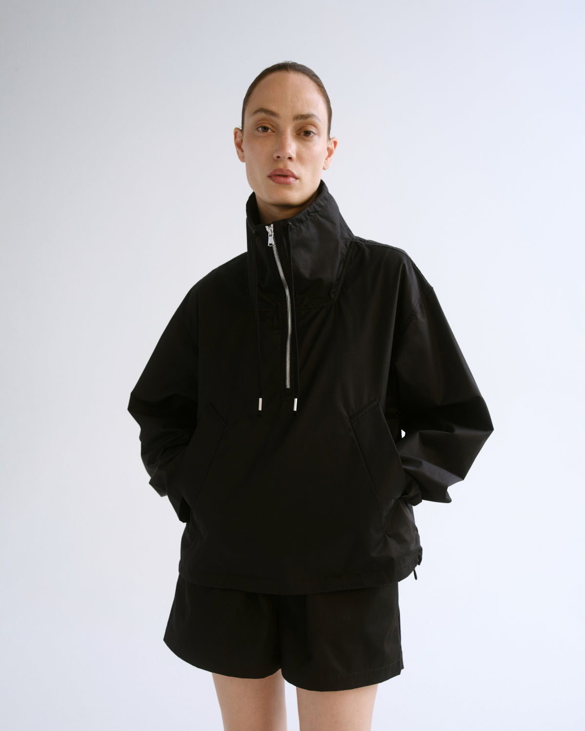 A model wearing a black high-collar zip-up jacket and matching black shorts, standing against a light gray studio background.
