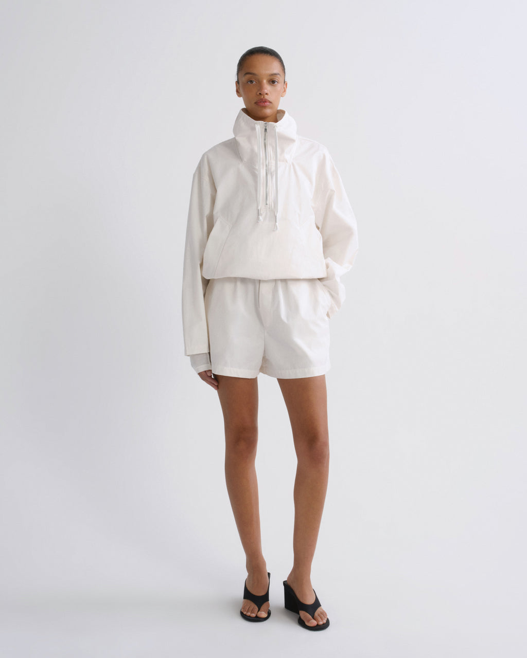 A model stands against a light gray studio background wearing a matching off-white cropped pullover jacket and shorts set, paired with black slide sandals.