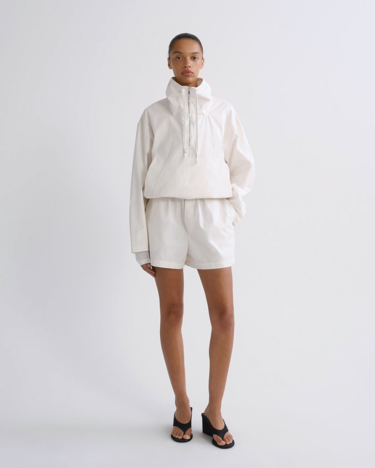 A model stands against a light gray studio background wearing a matching off-white cropped pullover jacket and shorts set, paired with black slide sandals.