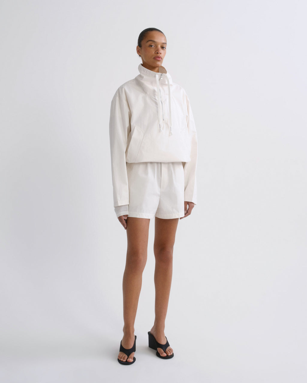 A model stands against a light gray studio background wearing a matching cream-colored two-piece outfit consisting of a cropped long-sleeve hoodie and shorts, paired with black slide sandals.