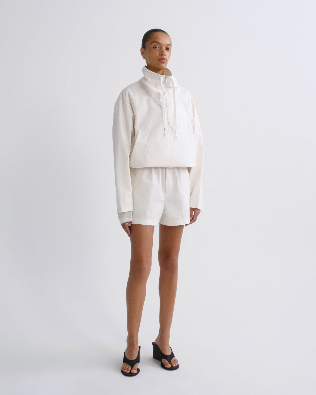 A model stands against a light gray studio background wearing a matching cream-colored two-piece outfit consisting of a cropped long-sleeve hoodie and shorts, paired with black slide sandals.