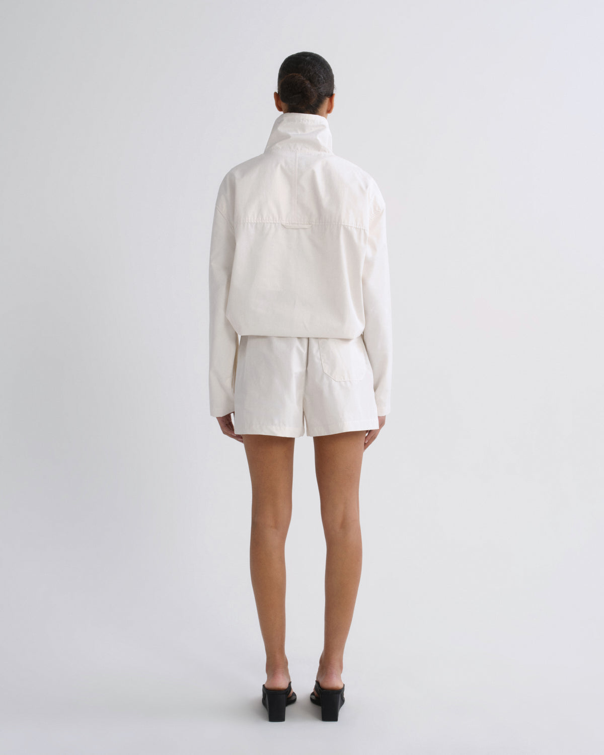 A model viewed from the back wearing a cream-colored cropped jacket and matching shorts, paired with black mule sandals, against a plain light gray studio background.