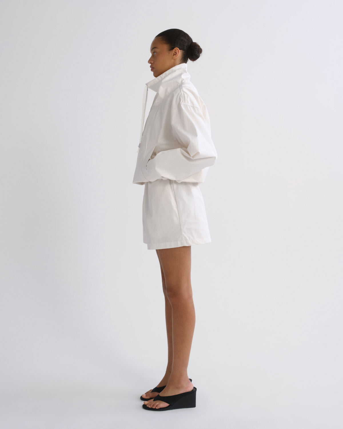 A model stands in profile wearing a white oversized cropped jacket with dramatic puffed sleeves and matching white shorts, paired with black wedge sandals, against a plain light gray studio background.