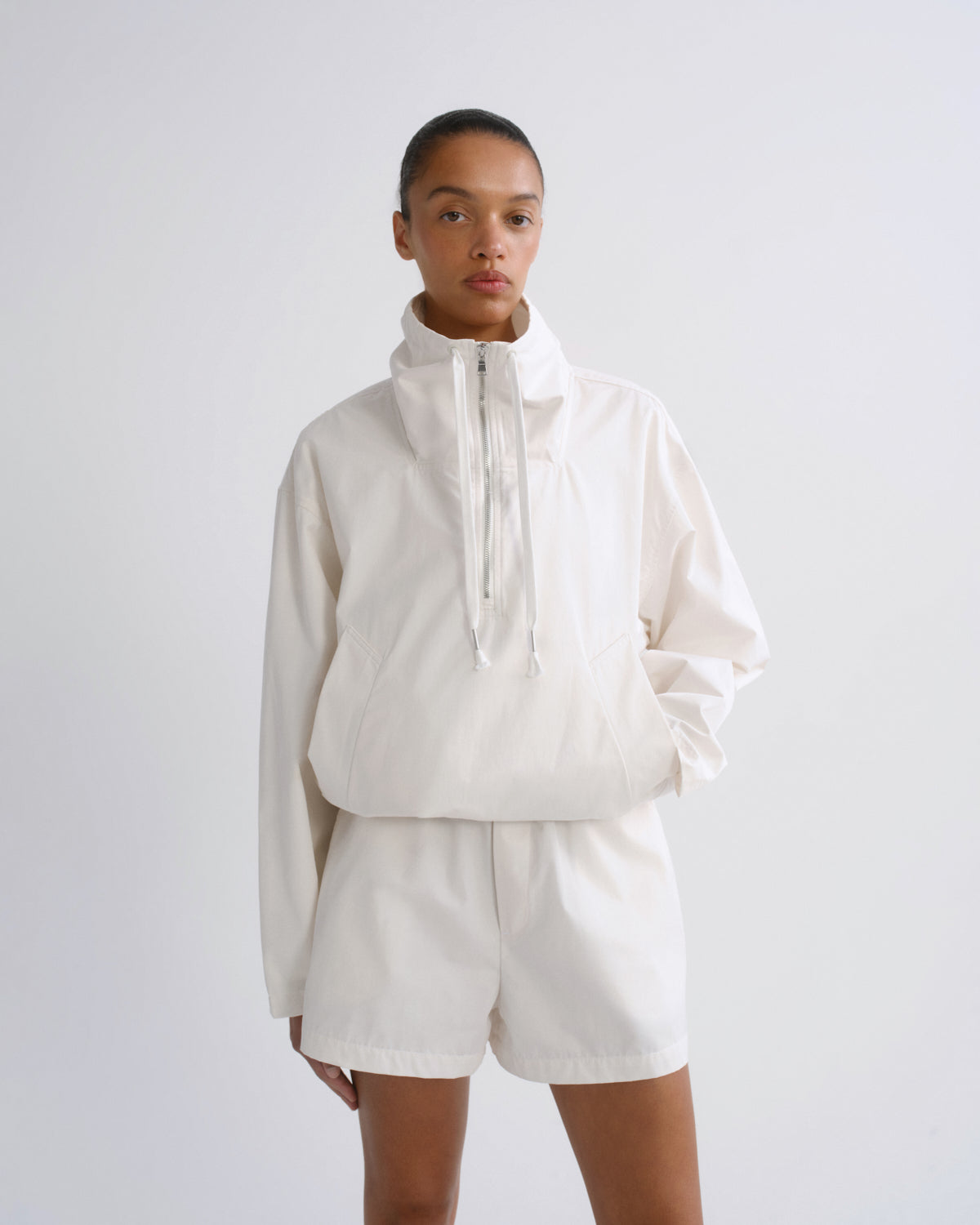 A model wearing a matching off-white cropped windbreaker jacket and shorts set, photographed against a seamless light gray studio background.