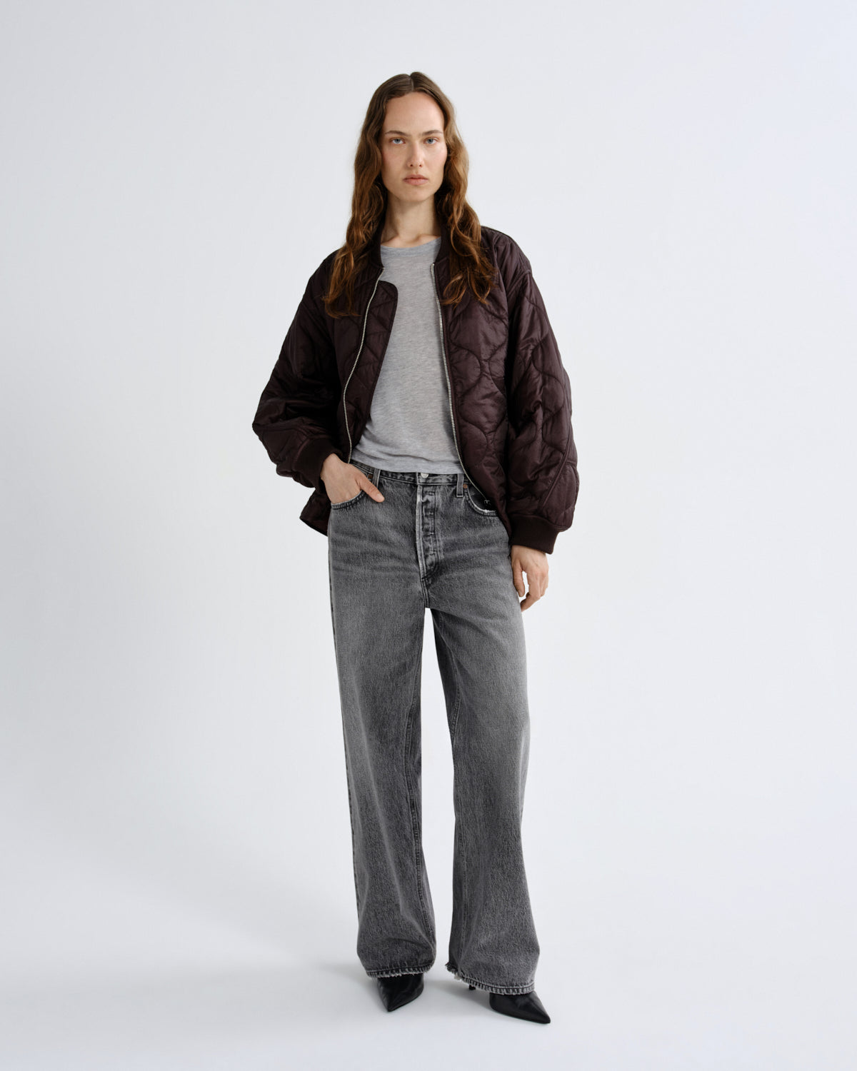 Person wearing a brown jacket, gray shirt, and black jeans on a white background