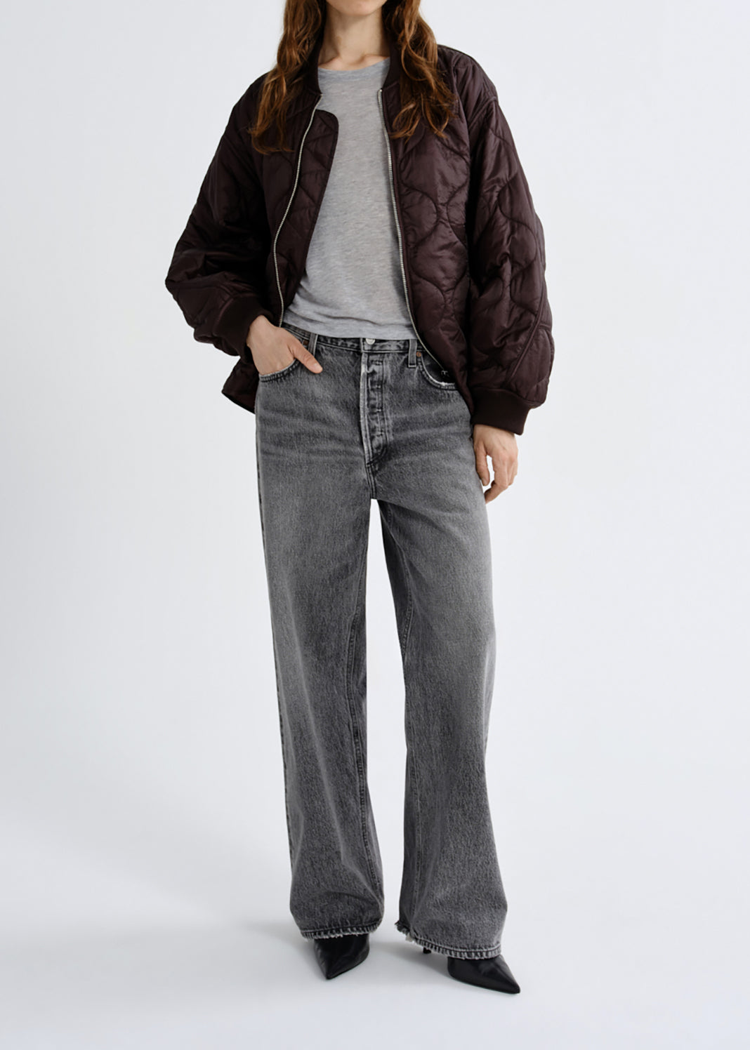 A model wearing a dark brown quilted bomber jacket, a grey crewneck t-shirt, wide-leg grey denim jeans, and black pointed-toe heels, photographed against a grey background.