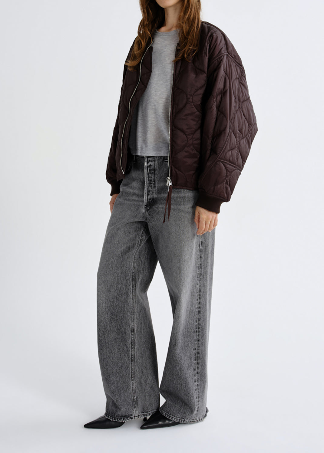A model wearing a dark brown quilted bomber jacket, a grey crewneck sweater, wide-leg grey jeans, and black pointed flats, photographed against a grey background.