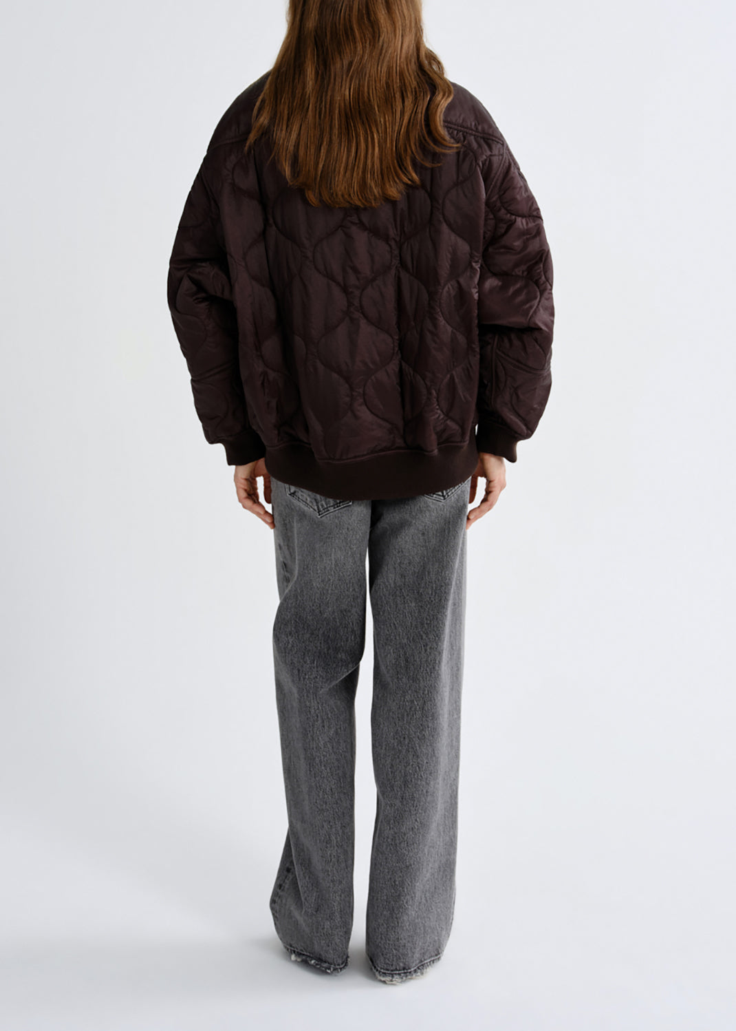 A person shown from the back wearing a dark brown quilted puffer jacket and wide-leg grey jeans against a grey background.