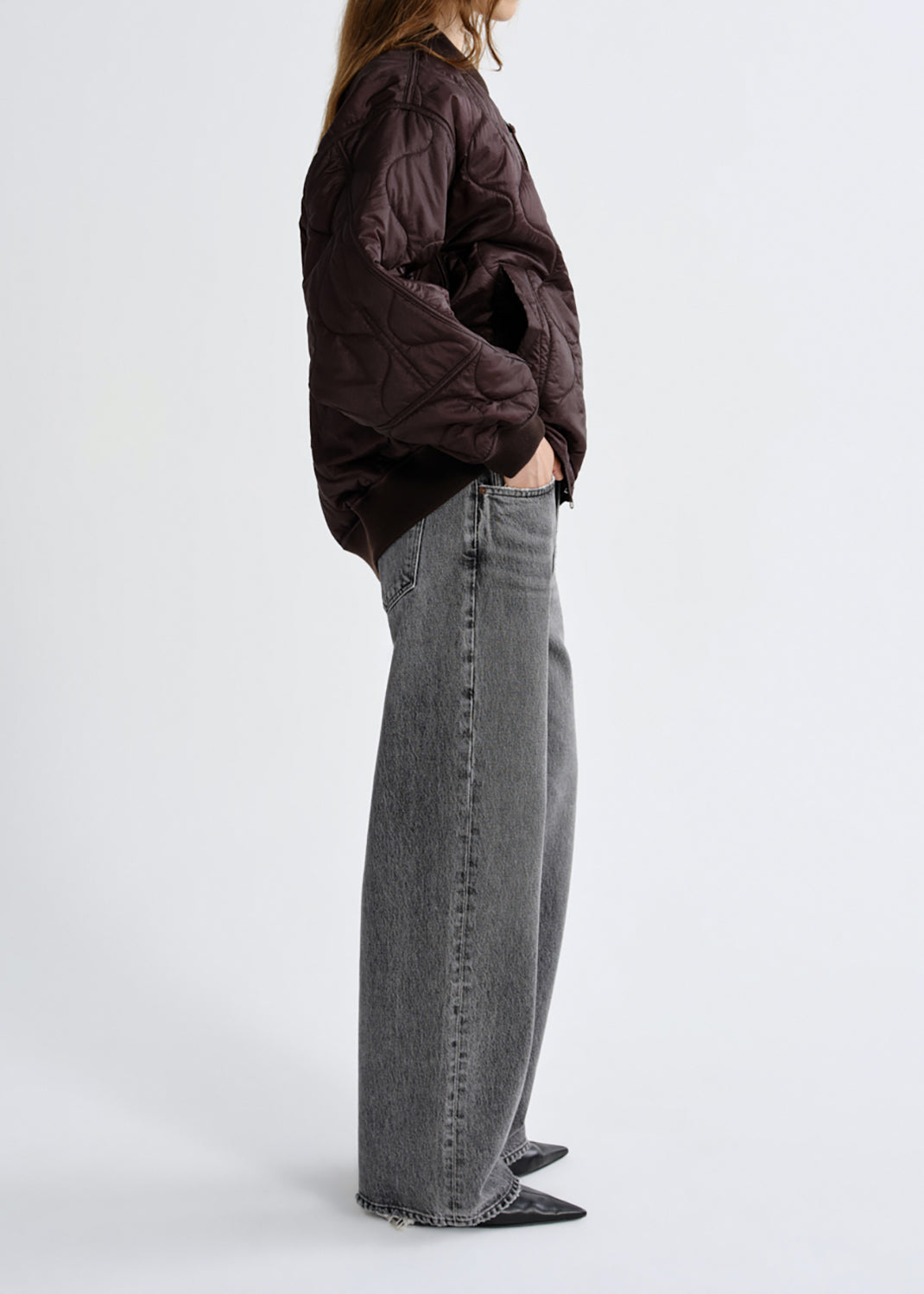 A model shown from the side wearing a dark brown quilted jacket and wide-leg grey jeans against a grey background.