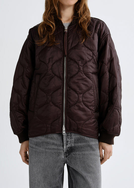 A person wearing a dark brown quilted zip-front jacket with a silver zipper, paired with grey jeans, against a grey background.